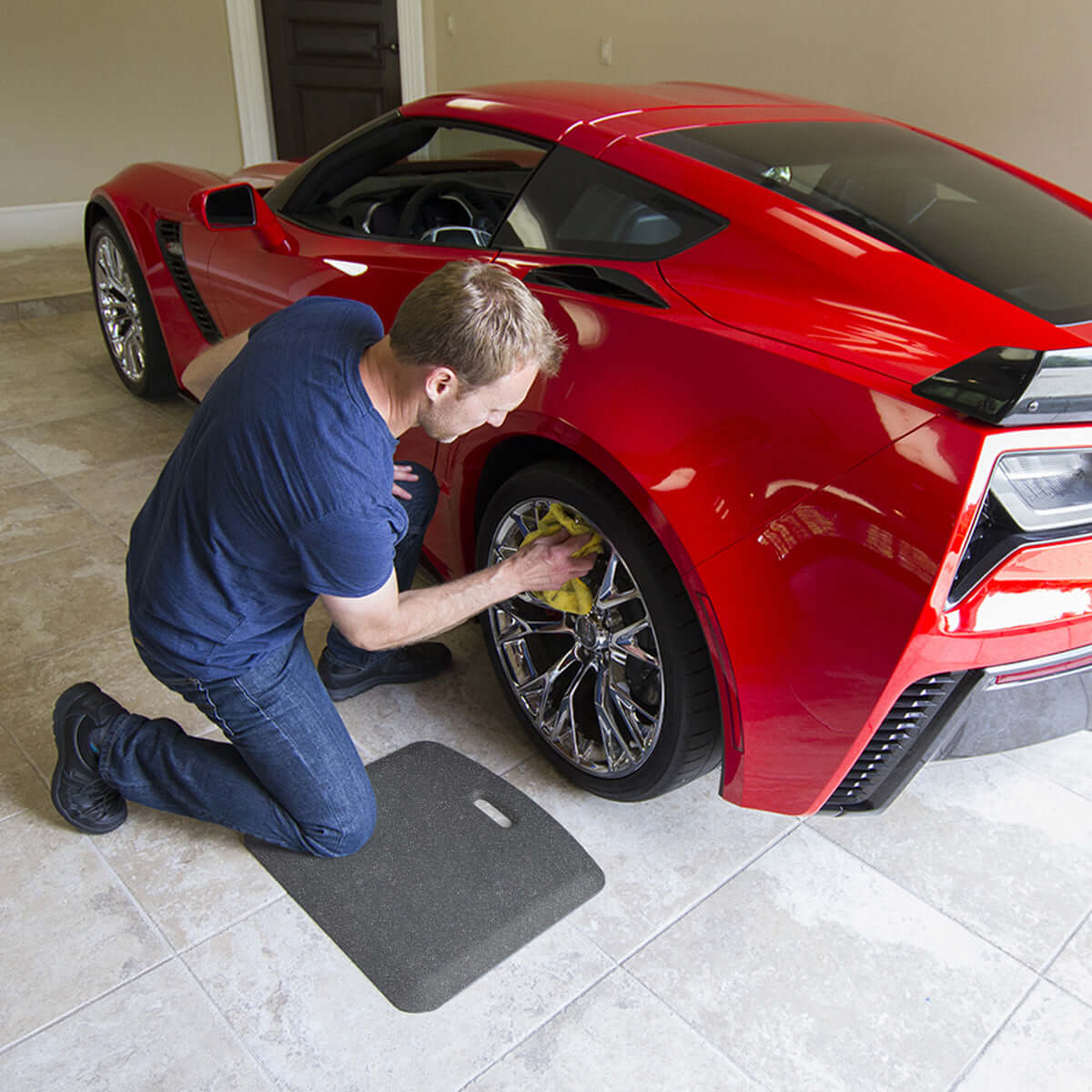 Man kneeling on a MobileMat while cleaning the wheel of a red sports car in a garage setting.